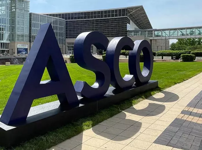 The image features large, bold letters spelling "ASCO" set against a green lawn. In the background, modern buildings with glass facades can be seen, along with walkways and trees, suggesting a bright and open outdoor environment.
