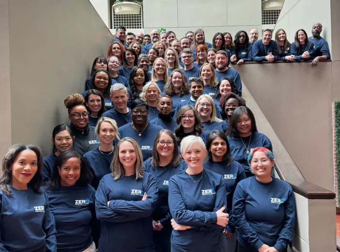 A large group of people standing on a staircase, all wearing matching navy blue shirts with the text "Bigger, Bolder, Zero" printed on them. They are smiling and posing for the photo in a well-lit indoor setting.