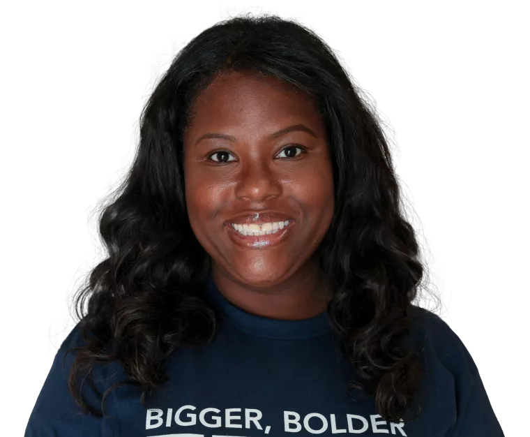 A person with long, dark wavy hair and a bright smile, wearing a navy blue sweatshirt with the words "BIGGER, BOLDER ZERO" in white