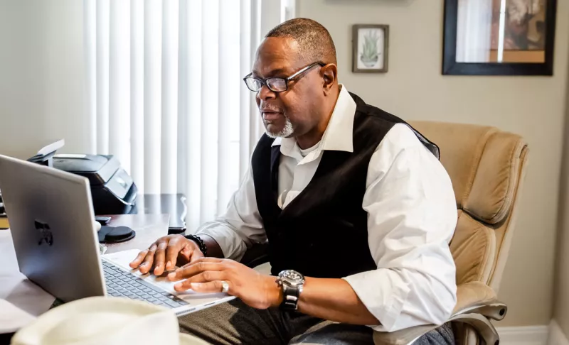 A man sits at a table with his laptop open, tuning into a virtual education session.