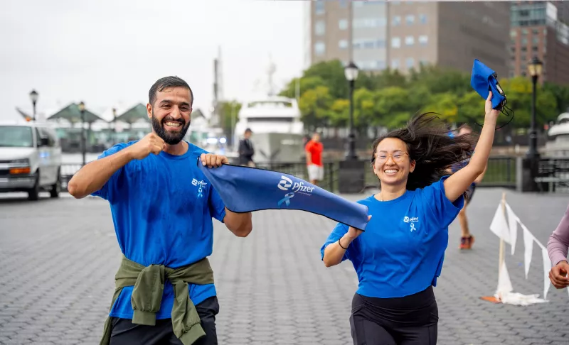 A man and a woman running joyfully, holding a blue Pfizer banner. They are wearing blue shirts, and the background shows a waterfront area with buildings and trees.
