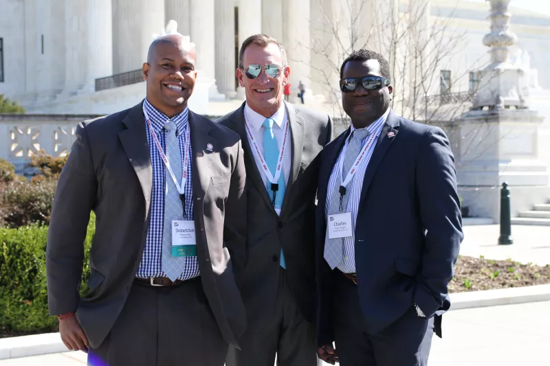 Three sharply dressed men in suits wearing lanyards and stand in front of a formal marble building