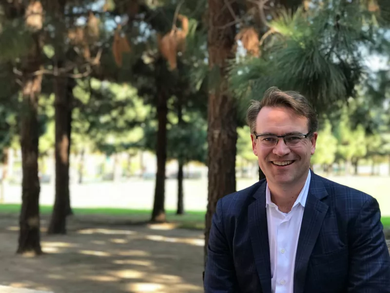 A man wearing smart looking glasses in a suit without a tie sits in shady trees out in the sun