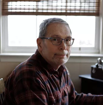 Older white male in flannel on and glasses sitting quietly for a headshot