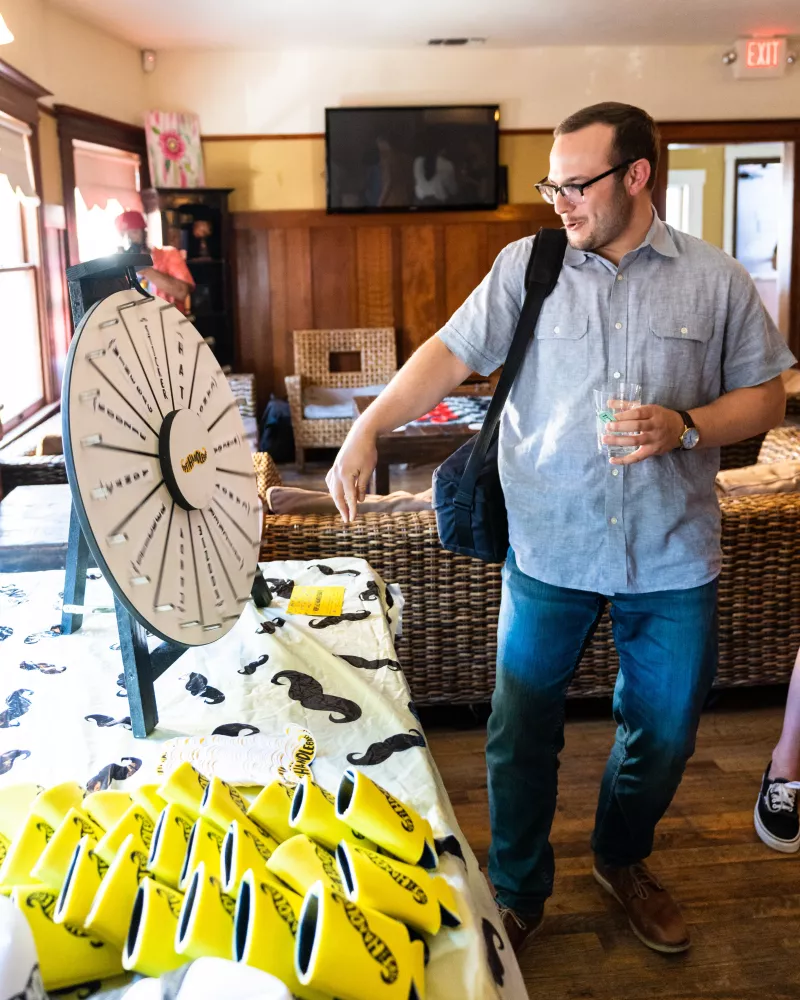 Man at an event stands in front of prize wheel and selection of mustaches and cozies 