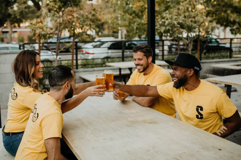 Four friends clink beers in yellow shirts at a table