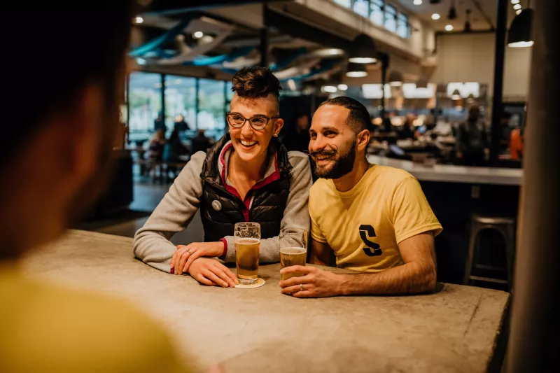 Two people sitting at a bar sharing beers smiling