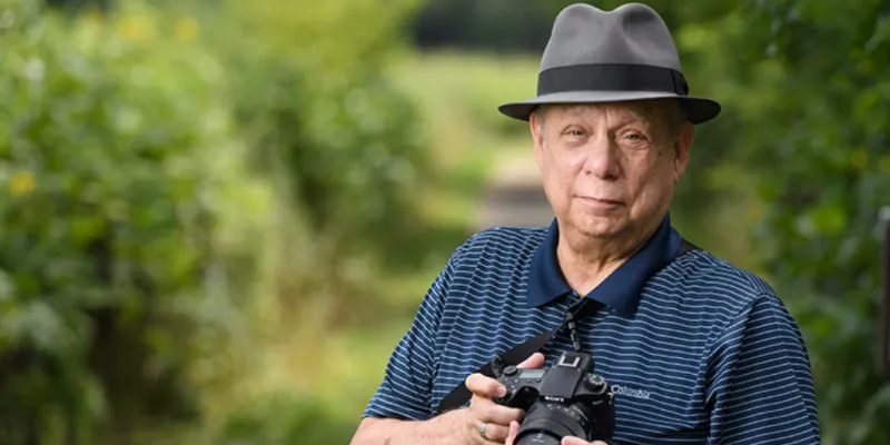 Older man wearing gray fedora holding a dslr camera on a forest path
