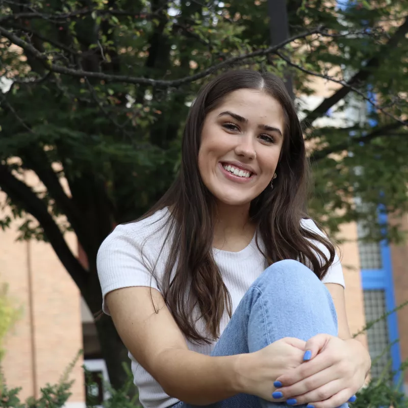 Brunette with long hair sitting in front of a dark green tree