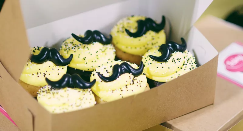 Yellow cupcakes decorated with black icing mustaches and black sprinkles sitting in a cardboard box