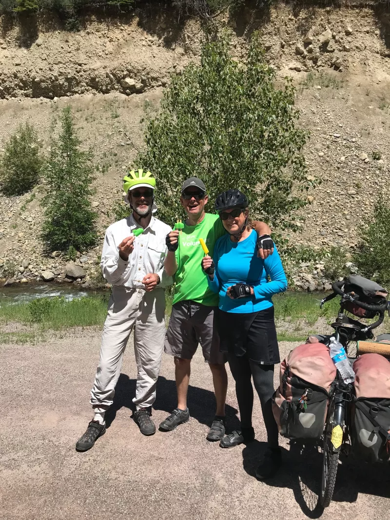 3 bicyclists pose by a mountainside holding up a trinket
