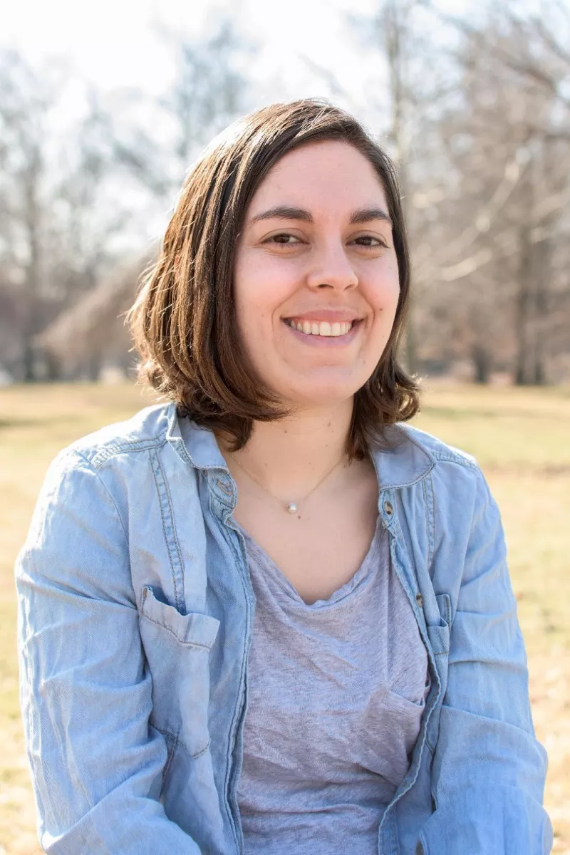 Smiling woman with mid length brown hair, a single pearl necklace and a light denim overshirt