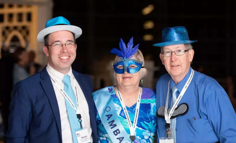 Three smiling people wearing bold and bright shades of blue, hats and one in a masquerade hat