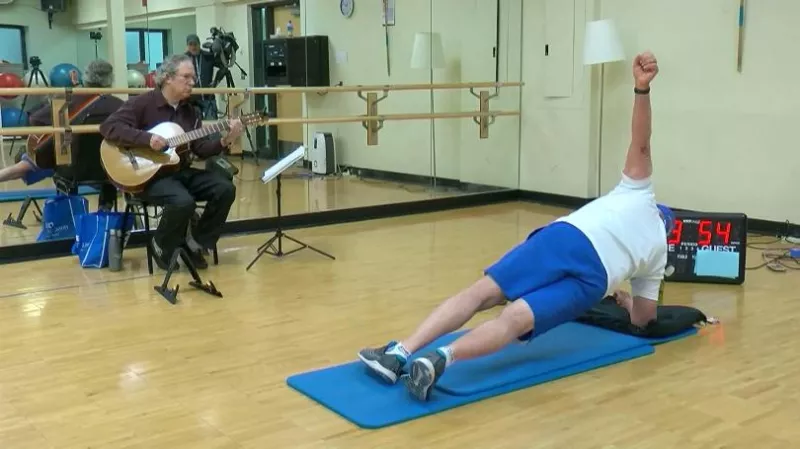 A man doing planks in front of another man playing a guitar