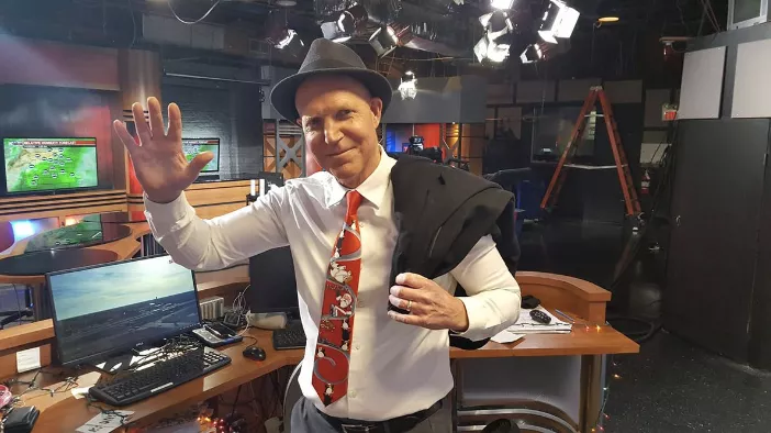 A man with a hat and a vest waving at the camera