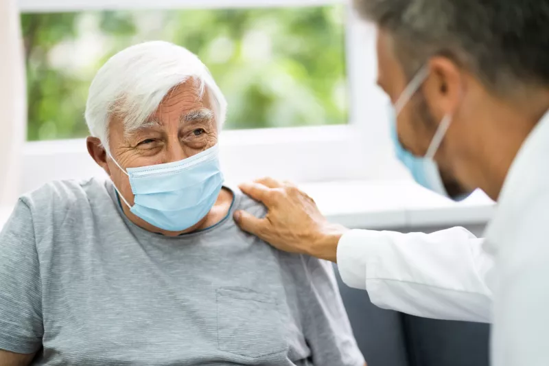 Elderly man with grey hair wearing a face mask and being comforted by his doctor