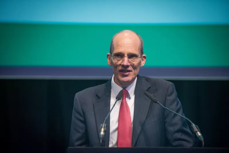 Bolding man in a suit and red tie giving a speech