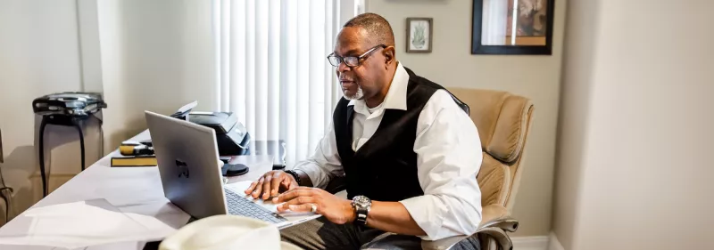 A man sits at a table with his laptop open, tuning into a virtual education session.