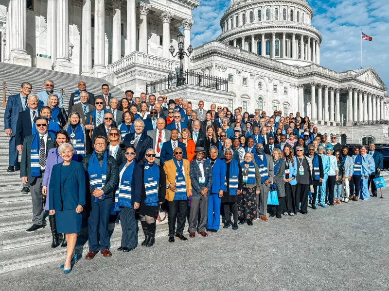 A large group of advocates stands on the steps of the U.S. Capitol, wearing blue attire and scarves with "ZERO" logos, symbolizing their support for prostate cancer awareness.