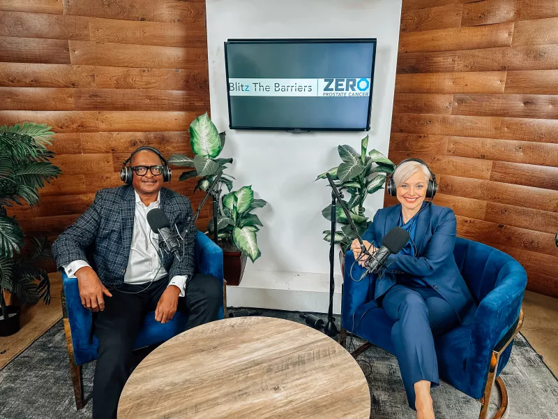 Two individuals are seated in a modern podcast studio. The man on the left wears glasses and a patterned blazer, looking relaxed. The woman on the right is in a navy suit with short platinum blonde hair, smiling warmly. Both wear headphones, ready for recording. A wooden wall and plants add a rustic touch, with a screen displaying "Blitz The Barriers" and "ZERO Prostate Cancer" in the background.