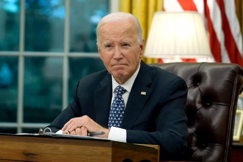 Former President Joe Biden in a suit sits at a desk in a formal office, looking serious. An American flag and lamp are in the background.