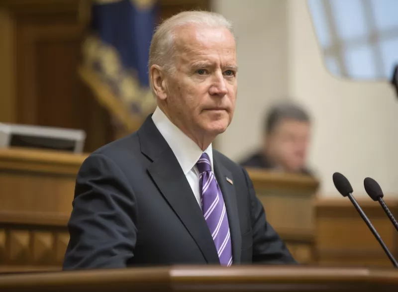Former President Joe Biden in a dark suit and purple striped tie stands at a podium, delivering a speech in a formal government setting. The background shows a blurred audience and ceremonial flags, indicating a high-level official event.