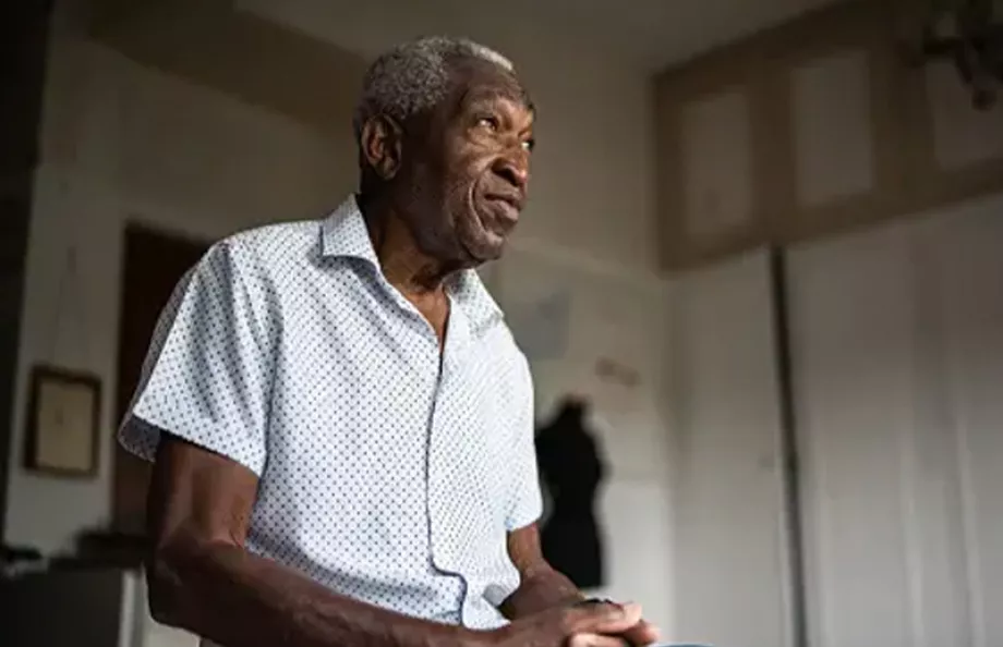 A Black man sitting on the edge of his bed with his hands together