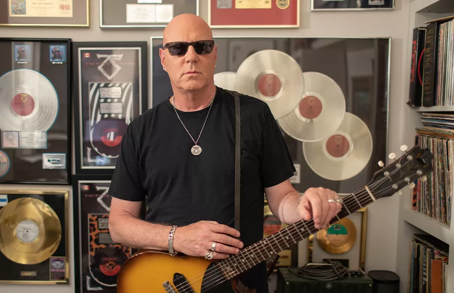 Jay Jay French holding a guitar standing in front of a wall with Twisted Sister records