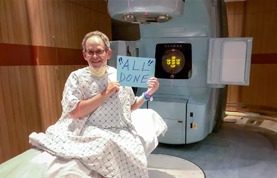 A senior man, Bob Jacobs, sitting in a hospital room with a radiation machine behind him. He's wearing a medical gown and holding a sign that says "All Done"