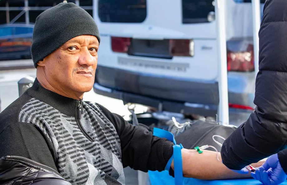 A black man wearing a beanie and long sleeves with his left sleeve rolled up. There is a tourniquet on his arm and he is getting his blood drawn.