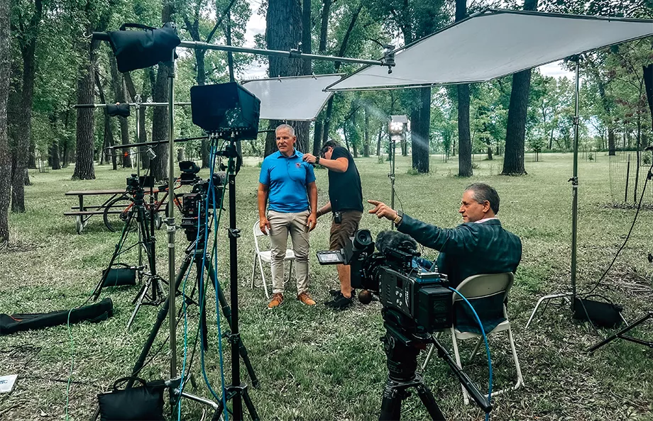 An older man in khaki pants and a blue shirt standing with lights and media all around him