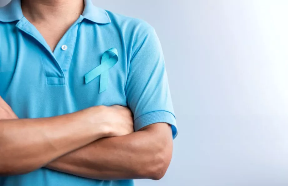 A person in a blue polo with a prostate cancer ribbon on the front. Their arms are crossed. 