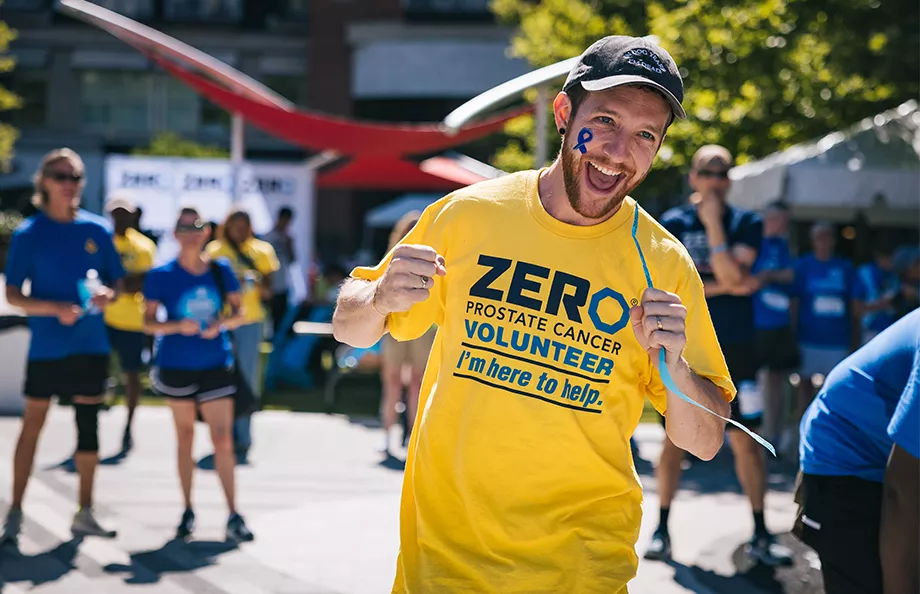 An energetic volunteer wearing a yellow ZERO Prostate Cancer Volunteer t-shirt with "I'm here to help" text smiles broadly at the camera on a sunny day. He has a blue ribbon painted on his face, with other volunteers visible in the background.