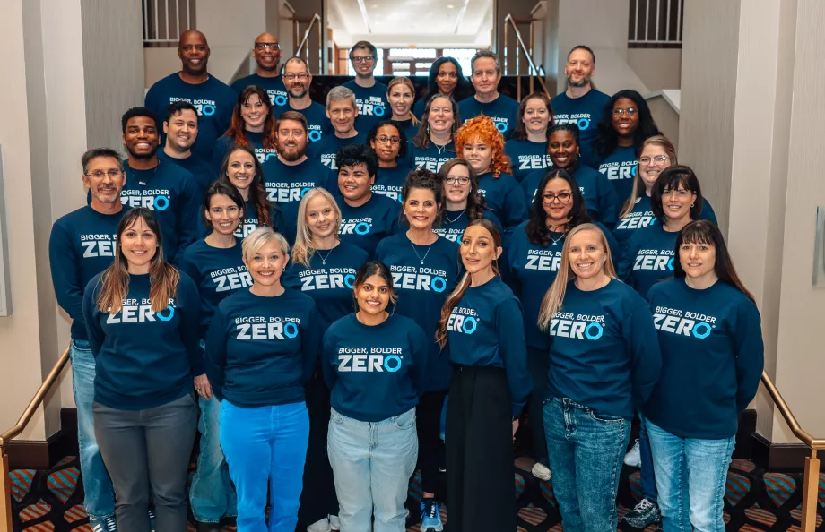 A large group of people standing on a staircase, all wearing matching navy blue shirts with the text "Bigger, Bolder, Zero" printed on them. They are smiling and posing for the photo in a well-lit indoor setting.