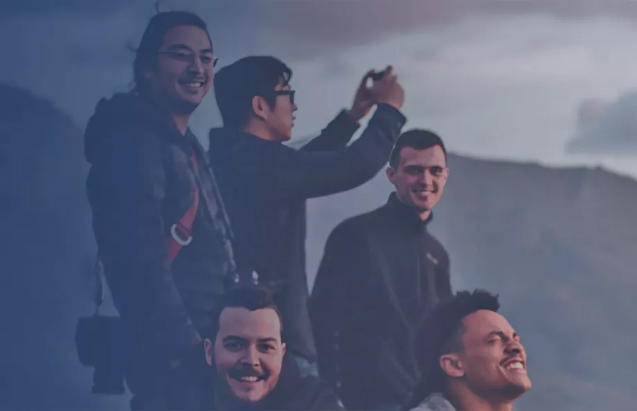 A group of five individuals enjoying an outdoor moment against a mountainous backdrop. The sky is partially cloudy, providing soft lighting. The leftmost person wears a dark jacket with a hood and holds a camera. The second person, slightly behind, is taking a photo with a device. The third person in the middle smiles warmly. The fourth and fifth individuals are also part of the group, sharing the experience.