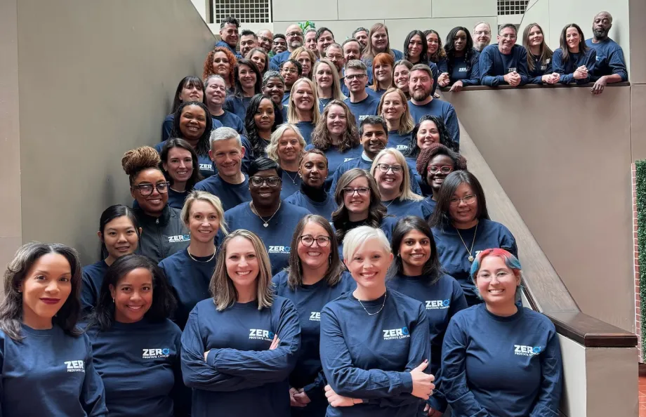A large group of people standing on a staircase, all wearing matching navy blue shirts with the text "Bigger, Bolder, Zero" printed on them. They are smiling and posing for the photo in a well-lit indoor setting.